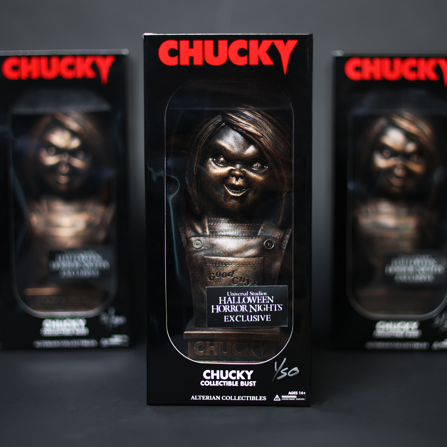 Front view of the Chucky Bronze Bust figure displayed with blurred packaged figures in the background.